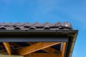 Close-up of dark graphite roof tiles, black aluminium guttering on brackets, wood trusses, blue sky.