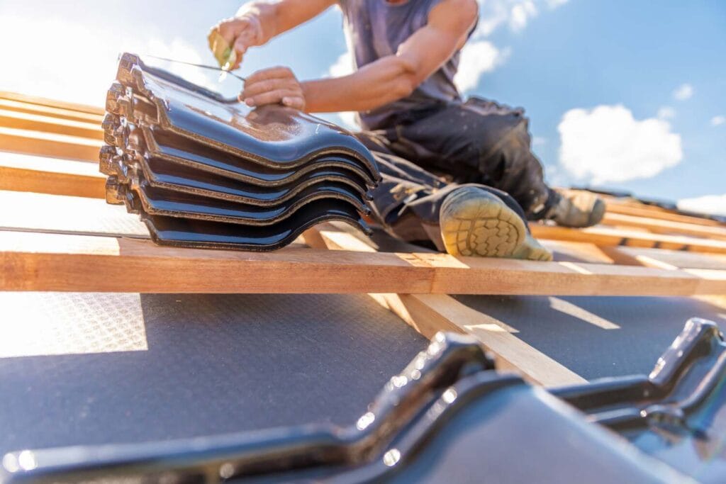 Roofer sitting on timber battens fixing glossy black ceramic roof tiles over a dark underlay membrane against blue sky.