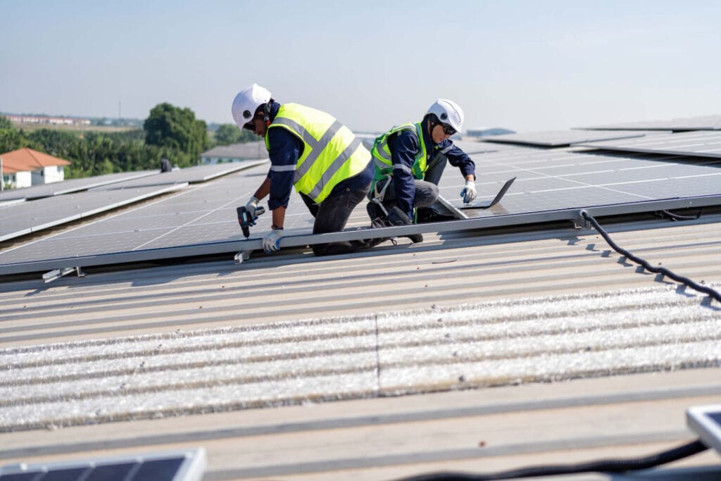 Two engineers in hi-vis vests and white hard hats drilling solar panel mounting rails on a large metal roof.